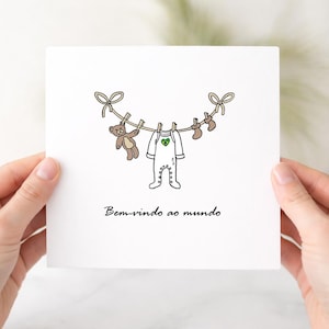 Puede incluir: Una tarjeta de felicitación blanca con un dibujo lineal de un tendedero con un oso de peluche marrón, un body blanco con un pequeño emblema de corazón verde y amarillo, y dos calcetines pequeños. El texto "Bem-vindo ao mundo" está en negro.