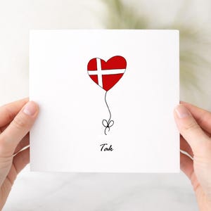 May include: A square white greeting card featuring a heart-shaped balloon with the red and white Danish flag pattern. A thin black line extends from the heart to a small bow at the bottom. The word Tak is printed in black script below the bow.
