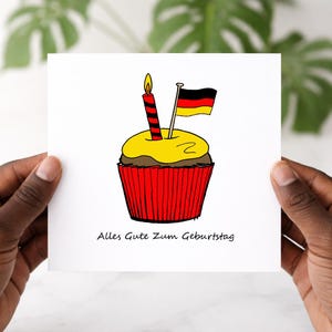 May include: A white card with a hand-drawn cupcake design. The cupcake has a candle and a German flag. The cupcake has a yellow top and a red liner. The text "Alles Gute Zum Geburtstag" is below.