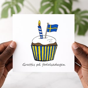 May include: A white greeting card featuring a cupcake illustration with a blue and yellow striped wrapper. The cupcake has white frosting, a blue candle, and a Swedish flag. The text "Grattis på födelsedagen" is written at the bottom.