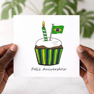 May include: A birthday card with a cupcake illustration, a lit candle, and a Brazilian flag. The cupcake has white frosting and a green, yellow, and blue striped wrapper. The text "Feliz Aniversário" is written below the cupcake.