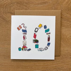 Puede incluir: Tarjeta blanca con el número "10" formado por ilustraciones coloridas de libros y lectores electrónicos. La tarjeta está sobre un sobre marrón, ideal para un cumpleaños temático de libros.