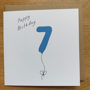 Peut inclure: Une carte blanche avec les mots "Happy Birthday" écrits à l'encre noire. Un ballon bleu en forme de chiffre 7 est dessiné sur la carte avec une ficelle noire et un nœud.