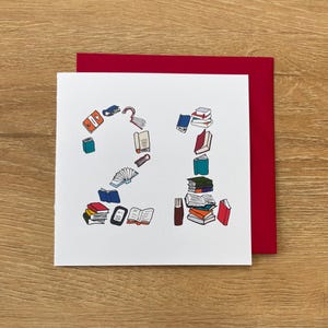 May include: A white card featuring the number "21" created from colourful book illustrations, a bookmark, and an e-reader. The card sits on a wooden surface with a red envelope in the background.