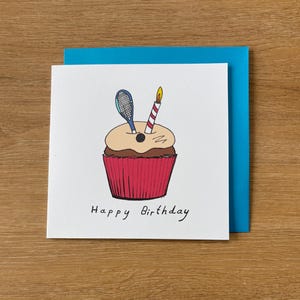Könnte beinhalten: Geburtstagskarte mit einer handgezeichneten Illustration eines Cupcakes mit Tennisschläger und Kerze. Der Cupcake hat eine rote Hülle, hellbraunes Frosting und die Worte "Happy Birthday" sind darunter geschrieben.