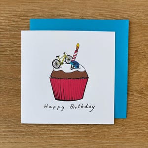 May include: A white birthday card with a blue envelope. The card shows a cupcake illustration with a bicycle, helmet, and lit candle. The words "Happy Birthday" are written below the cupcake.