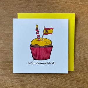 Puede incluir: Tarjeta de felicitación de cumpleaños con una ilustración de cupcake, una vela y una bandera española. El cupcake tiene una parte superior amarilla y un envoltorio rojo. Las palabras "Feliz Cumpleaños" están impresas debajo.