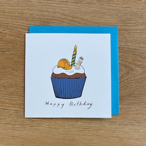 May include: A birthday card with a cupcake illustration, designed with a construction theme. The cupcake has a blue liner, chocolate frosting, and is topped with a yellow hard hat, a candle, and a blueprint. The words "Happy Birthday" are written below.