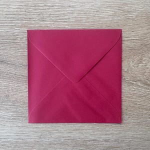 May include: A square, magenta-colored envelope with a pointed flap, resting on a light wood-grain surface. The envelope's color is a deep, rich shade of red-purple. The flap is folded over, creating a triangular shape.