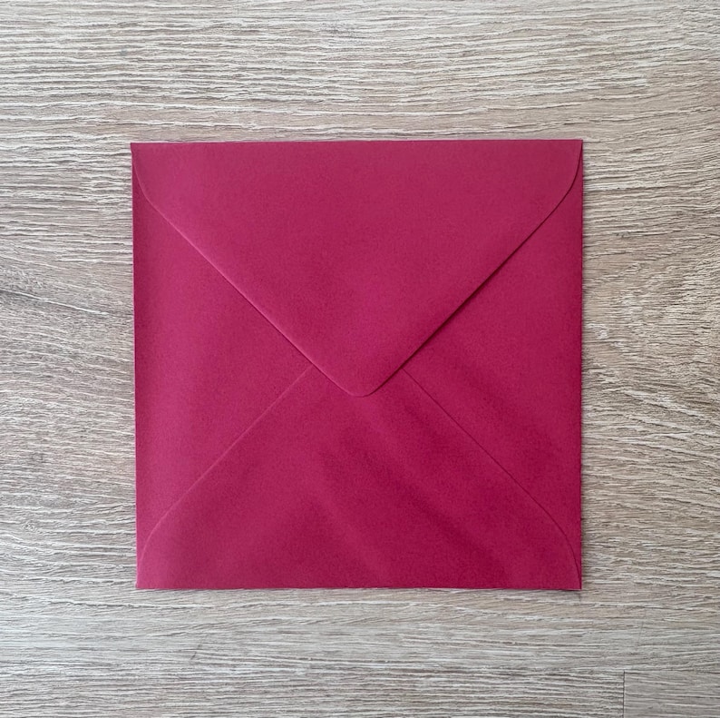 May include: A square, magenta-colored envelope with a pointed flap, resting on a light wood-grain surface. The envelope's color is a deep, rich shade of red-purple. The flap is folded over, creating a triangular shape.