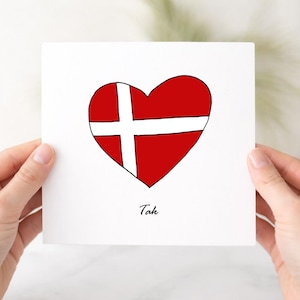 May include: A square white greeting card featuring a hand-drawn red heart with a white cross pattern resembling the Danish flag. Below the heart, the word Tak is printed in a simple black script font. The design is centered on the card.