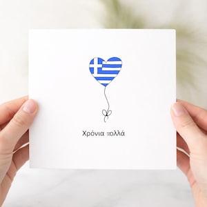 May include: A square white greeting card featuring a heart-shaped balloon with the blue and white Greek flag pattern. A thin black string with a small bow hangs from the heart. The Greek text "Χρόνια πολλά" is printed in black below the balloon.