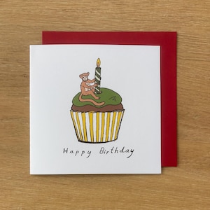 May include: A square white greeting card featuring a hand-drawn illustration of a brown monkey sitting on a cupcake with green frosting and a striped candle. The cupcake has a yellow and white striped liner. The text Happy Birthday is below.