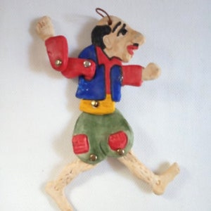 May include: A whimsical handmade figurine of a person with a red, blue, and yellow outfit. The figurine is made of clay and has a painted face with a large nose. The figurine is posed in a running position with one arm raised.