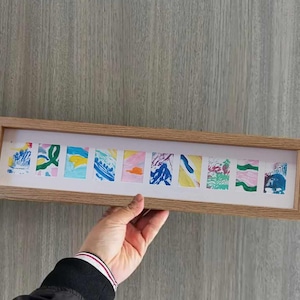 Può includere: Una cornice in legno con 10 piccoli dipinti astratti colorati al suo interno. I dipinti sono di vari colori, tra cui blu, verde, giallo, rosa e arancione.