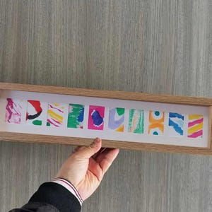 Può includere: Una cornice in legno con 10 dipinti astratti colorati al suo interno. I dipinti sono di diverse dimensioni e forme e sono disposti in fila. La cornice è di colore marrone chiaro e ha un passepartout bianco.