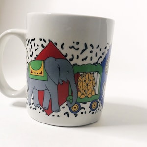 Puede incluir: Taza de cerámica blanca con un diseño de dibujos animados colorido que presenta un elefante, un león en una jaula y una carpa roja. El diseño está sobre un fondo blanco con formas geométricas en blanco y negro.