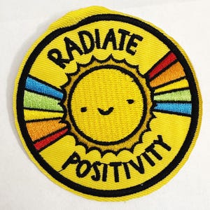 Puede incluir: Parche bordado amarillo con un diseño de sol sonriente y las palabras "RADIATE POSITIVITY" en negro. El sol tiene rayos de arcoíris. El parche es circular con un borde negro.