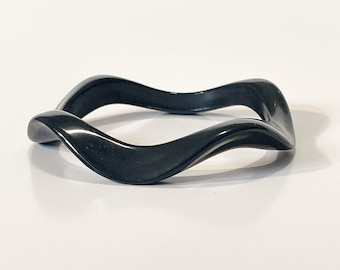 May include: A black, wave-shaped bangle. The bracelet has a smooth, glossy finish and a distinctive, undulating design. The bracelet is made of a solid, dark material and is displayed on a white surface.