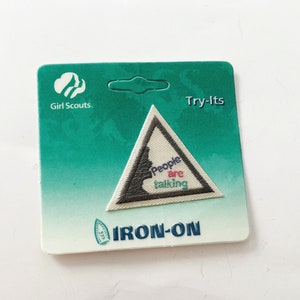 May include: A white triangular Girl Scout patch with a gray silhouette of a person talking. The patch has the text "People are talking" and "Try-Its" and "IRON-ON" printed on it.