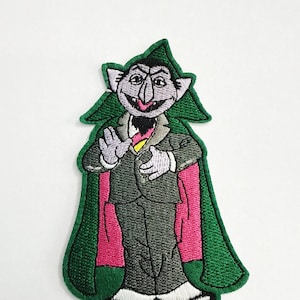 May include: Embroidered patch featuring a cartoon character, The Count, from Sesame Street. The patch depicts The Count in a dark gray suit, green cape, and pink lining. The Count is smiling and has white gloves. The patch has a green border.