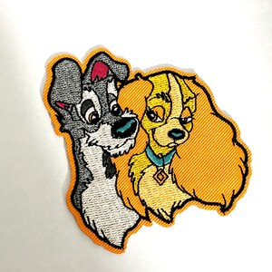 Op de afbeelding: Geborduurde patch met de Disney-personages Tramp en Lady uit de film "Lady en de Vagebond". De patch heeft een oranje achtergrond. Zwerver is grijs en wit, en Lady is geel met een blauwe halsband en een ruitvormige hanger.