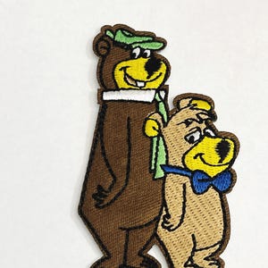 May include: Embroidered patch featuring two cartoon bears. One bear is brown with a green hat, white collar, and green scarf. The other bear is tan with a blue bow tie and a yellow hat. The patch has a brown border.