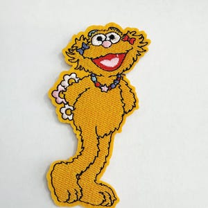 Puede incluir: Parche bordado con un personaje de dibujos animados amarillo con una gran sonrisa, que lleva un collar de flores y accesorios para el pelo. El parche tiene un contorno negro sobre un fondo blanco. El personaje está de pie.
