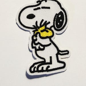 Puede incluir: Un parche de tela bordado que muestra un perro de dibujos animados blanco con contornos negros, una oreja negra y una nariz negra. El perro sostiene un pequeño pájaro amarillo brillante entre sus patas, con la cabeza del pájaro visible cerca.