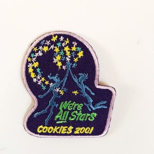 Op de afbeelding: Een paarse geborduurde patch met twee figuren die naar een uitbarsting van kleurrijke sterren reiken. De tekst "We're All Stars" en "COOKIES 2001" zijn geborduurd in groen en geel.