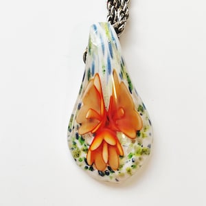 May include: A teardrop-shaped glass pendant necklace with a silver chain. The pendant features a colorful, abstract design with a central orange flower.