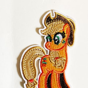 Peut inclure: Un patch brodé thermocollant représentant un poney de dessin animé avec des cheveux bruns, portant des lunettes et un chapeau brun. Le poney est orange et jaune avec une queue et une crinière brunes.