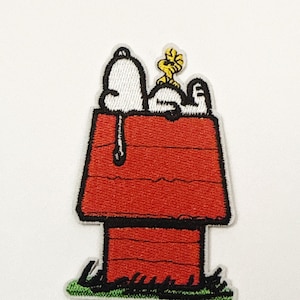 以下が含まれることがあります： スヌーピーとウッドストックがスヌーピーの赤い犬小屋の上にいる刺繍パッチ。パッチは黒い縁取りと下部に緑の草があります。スヌーピーは白く、ウッドストックは黄色です。