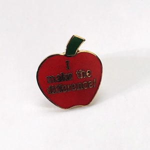 May include: Red apple-shaped enamel pin with a gold outline. The apple has a green stem and leaf. The text on the apple reads "I make the difference!"