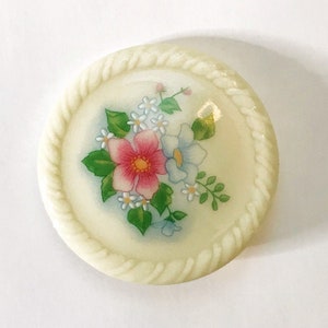 May include: A white ceramic button with a floral design. The button features a pink flower with blue and white flowers surrounding it. The button has a rope-like border.