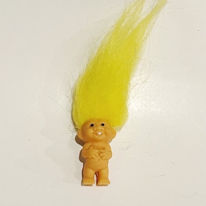 May include: A small, yellow-haired troll doll with a toothy grin. The troll is standing upright with its arms crossed in front of its chest.