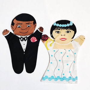 May include: A pair of hand puppets depicting a bride and groom. The bride is wearing a white dress with blue polka dots and a blue headband. The groom is wearing a black tuxedo with a white bow tie and a red rose.