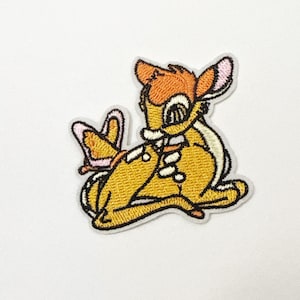 May include: Embroidered patch featuring a cartoon deer in shades of yellow, orange, and white. The deer is in a seated position with a butterfly nearby. The patch has a white border and is likely for clothing or accessories.