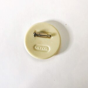 May include: White round pin with a silver pin back. The pin has the word "AVON" embossed on the back.
