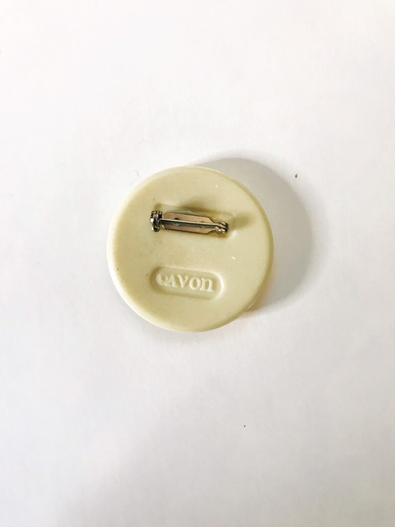 May include: White round pin with a silver pin back. The pin has the word "AVON" embossed on the back.