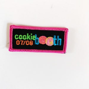 Op de afbeelding: Een geborduurde zwarte en roze patch met de tekst "cookie booth 07/08" en een kleurrijke afbeelding van drie koekjeswervelingen.