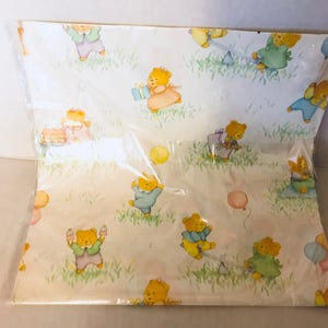 May include: A white wrapping paper with a repeating pattern of teddy bears wearing different colored clothes and holding balloons. The bears are surrounded by green grass and flowers.