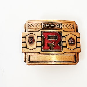 May include: A gold-colored metal belt buckle with a red and black letter "R" in the center. The buckle has a raised border and the year "1954" is inscribed above the letter "R". The buckle also has two smaller shields on either side of the letter "R", with the letters "B" and "A" inscribed in them.
