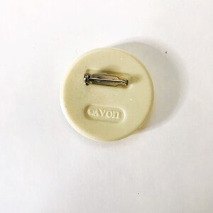May include: A white round pin with a silver pin back. The pin has the word "Cayon" printed on it.