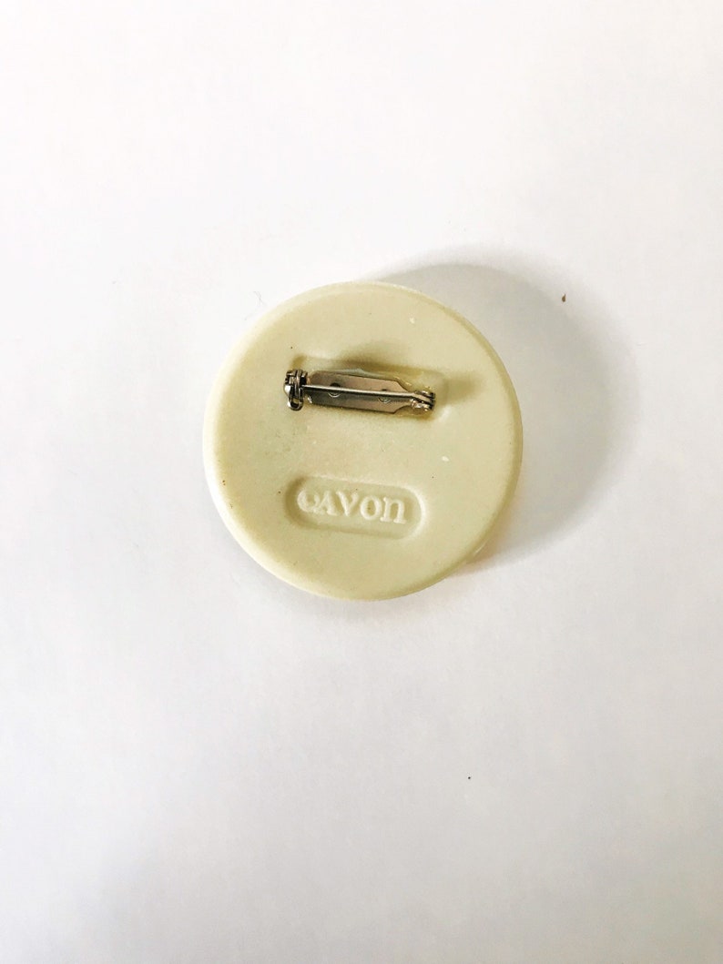 May include: A white round pin with a silver pin back. The pin has the word "Cayon" printed on it.