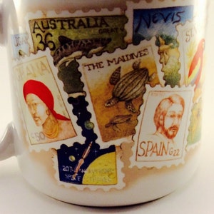 May include: A white ceramic mug with colorful postage stamps featuring images of a shark, a turtle, a person with a beard, and a person with a red headband. The stamps have text including "Australia 36", "The Maldives", "Spain G22", and "Nevis".
