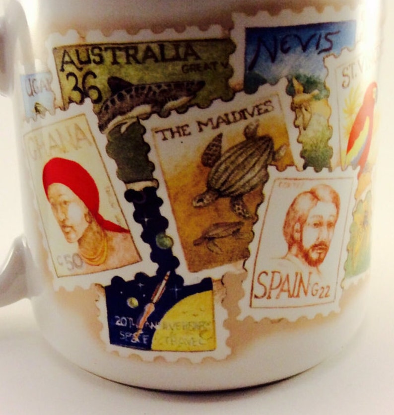 May include: A white ceramic mug with colorful postage stamps featuring images of a shark, a turtle, a person with a beard, and a person with a red headband. The stamps have text including "Australia 36", "The Maldives", "Spain G22", and "Nevis".