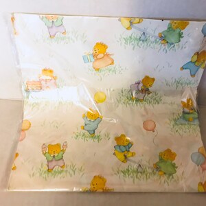 May include: A white wrapping paper with a repeating pattern of teddy bears wearing different colored clothes and holding balloons. The bears are surrounded by green grass and flowers.