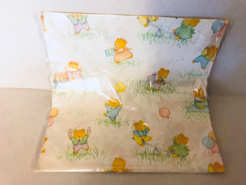 May include: A white wrapping paper with a repeating pattern of teddy bears wearing different colored clothes and holding balloons. The bears are surrounded by green grass and flowers.