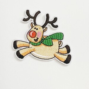 May include: Embroidered patch featuring a cartoon reindeer in mid-leap. The reindeer has a tan body, brown antlers, a red nose, and a green scarf. The patch is outlined in white.